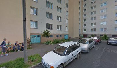 Lakóépület parkolója, emberek ülnek a padon.